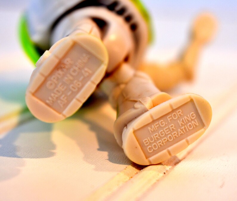 Może przedstawiać: Zbliżenie st&oacute;p figurki Buzz Lightyear. Podeszwy st&oacute;p są w kolorze beżowym z nadrukiem "MFG. FOR BURGER KING CORPORATION" i "&copy;PIXAR MADE IN CHINA AF-06-41". Figurka leży na białej powierzchni.