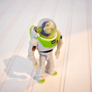 Może przedstawiać: Figurka akcji Buzz Lightyear, z białym i zielonym kombinezonem kosmicznym i przezroczystym hełmem. Zabawka ma szczeg&oacute;łowy design, w tym fioletową głowę i mały emblemat na klatce piersiowej. Figurka stoi na białej powierzchni.