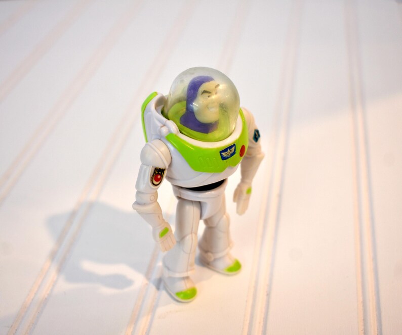 Może przedstawiać: Figurka akcji Buzz Lightyear, z białym i zielonym kombinezonem kosmicznym i przezroczystym hełmem. Zabawka ma szczeg&oacute;łowy design, w tym fioletową głowę i mały emblemat na klatce piersiowej. Figurka stoi na białej powierzchni.