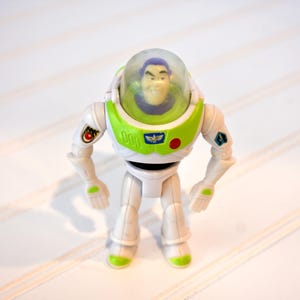 Może przedstawiać: Figurka akcji Buzz Lightyear, z białym i zielonym wzorem kombinezonu kosmicznego. Zabawka ma przezroczysty hełm, uśmiechniętą twarz i r&oacute;żne detale emblemat&oacute;w. Figurka stoi na białej powierzchni.