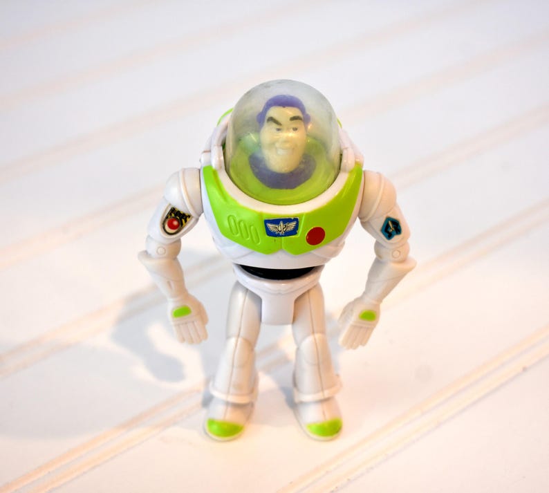 Może przedstawiać: Figurka akcji Buzz Lightyear, z białym i zielonym wzorem kombinezonu kosmicznego. Zabawka ma przezroczysty hełm, uśmiechniętą twarz i r&oacute;żne detale emblemat&oacute;w. Figurka stoi na białej powierzchni.