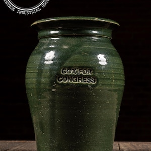May include: A vintage green ceramic crock with the embossed text "COX FOR CONGRESS". The crock has a rounded body, a slightly flared rim, and horizontal ridges. It is displayed on a wooden surface.