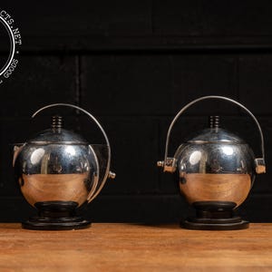 May include: Two silver, round, teapots with black bases and handles. The teapots are on a wooden surface.