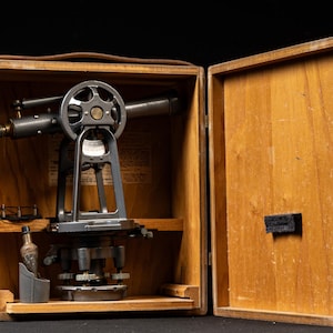 May include: A vintage metal surveying transit instrument housed inside an open wooden carrying case. The grey metal device features a telescope, a circular adjustment wheel, and a base with leveling screws. A small glass vial sits in a holder.