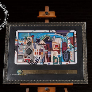 May include: Framed artwork featuring a colorful abstract cityscape with geometric shapes and buildings. The artwork is displayed on a wooden easel. The frame is dark with a metallic finish. The artwork is labeled "Millennium Graphics."