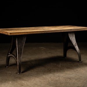 May include: A rustic wooden table top with a dark brown finish sits atop two black cast iron industrial style legs. The table is approximately 6 feet long and 3 feet wide.