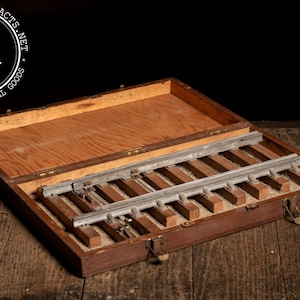 May include: Open wooden box with a hinged lid, containing metal and wooden components. The box is brown with a lighter wood interior. The metal pieces are silver. The box is on a wooden surface. The logo "INDUSTRIALARTIFACTS.NET" is in the upper left corner.