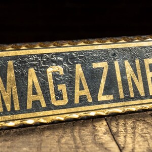 May include: Rectangular black sign with the word "MAGAZINES" in gold lettering. The sign has a gold-colored border and is set against a dark background. The sign is placed on a wooden surface.