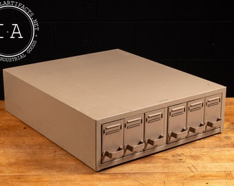 May include: A light gray metal drawer unit with six drawers. Each drawer has a metal handle and a slightly raised lip around the edge.