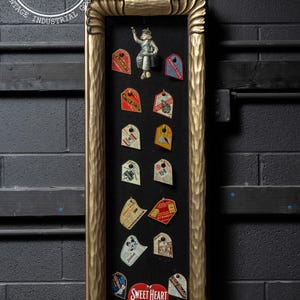 May include: A gold-framed display with a black background showcasing a collection of vintage advertising tags. The tags feature various products, including flour, cornmeal, and a heart-shaped tag for Sweet Heart Products.