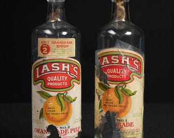 May include: Two vintage glass bottles of Lash's Quality Products orangeade. Each bottle has a label with an orange illustration and text. The labels are peeling, showing the age of the bottles. The bottles are empty.
