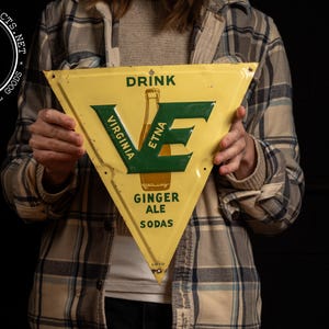 May include: Vintage triangular yellow metal sign for Virginia Etna Ginger Ale Sodas. The sign features green lettering and a bottle illustration. The sign is held by a person wearing a plaid shirt.
