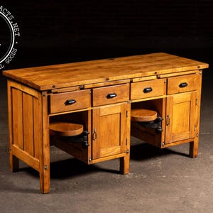 May include: A wooden desk with a warm, honey-toned finish. It features four drawers with black handles, two cabinets, and two pull-out circular shelves. The desk is sturdy and has a vintage, industrial aesthetic.