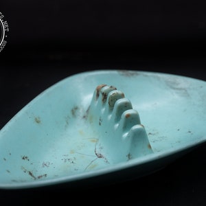 May include: A light blue ceramic ashtray with a worn, vintage look. The ashtray has a unique, triangular shape with a raised center section for holding cigarettes.