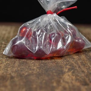 May include: A clear plastic bag tied with a red string, filled with several red, translucent candies. The bag sits on a textured, brown wooden surface, with a dark background.