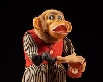 May include: A vintage wind-up toy monkey wearing a red vest and black and white striped pants. The monkey is holding a red cup with dice on it. The monkey has blue eyes and a brown fur coat.