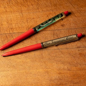 May include: Two red pens with clear plastic barrels. One pen has a cityscape inside and the other says "Shaw International Airport".