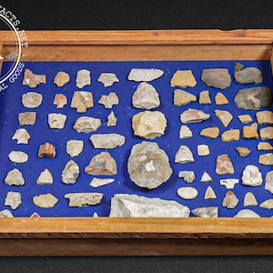 May include: A collection of various shaped stones displayed on a blue fabric within a wooden framed box. The stones are in shades of gray, tan, and brown. The box has the text "INDUSTRIAL ARTIFACTS" and "IA" in a circle.