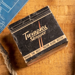 May include: A black box with the words "Twinplex Stropper" and "Makes the Best Razor Blade Better" printed on it. The box is sitting on a wooden surface.
