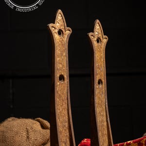 May include: Two antique, ornate, gold-toned metal brackets with a distressed finish. Each bracket has a decorative top and a central hole. The background is dark, with a burlap sack and a red and gold patterned cloth visible.
