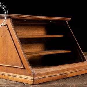 May include: A vintage wooden display case with a slanted glass front and two shelves. The case is made of stained wood and sits on a wooden surface. The display case is empty and ready to be filled with collectibles.
