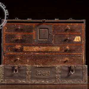 May include: A weathered wooden tool chest featuring multiple drawers of varying sizes, a central cabinet door, and a dark metal base with two front latches. The metal base displays the embossed letters D.S.H. and shows signs of oxidation and wear.