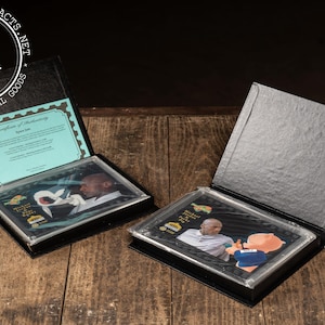 May include: Two black display cases with images of a man, one open and one closed. The open case reveals a certificate of authenticity. The cases are on a wooden surface. The Industrial Artifacts logo is in the upper left corner.