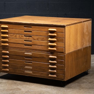 May include: A large wooden flat file cabinet featuring two stacked sections with a total of ten drawers. Each drawer has a horizontal wooden handle on the right side. The cabinet is supported by thin black metal legs and has a flat top surface.