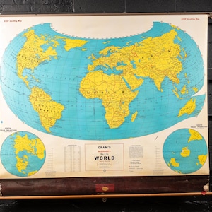 May include: A vintage world map with a blue ocean and yellow continents, labeled "Cram's Beginner's World." The map includes polar projections and is mounted on a wooden roller. The map is hanging on a black brick wall.