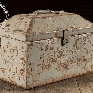 May include: Vintage metal toolbox with a hinged lid and a handle. The box is painted a faded gray color with areas of rust and wear, giving it a distressed appearance. The toolbox is rectangular and sits on a wooden surface.