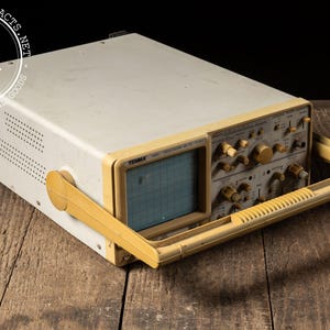May include: Vintage beige and yellow oscilloscope with a handle. The device has a screen with a grid pattern and numerous knobs and dials. The word "TENMA" is visible on the front. The device is on a wooden surface.