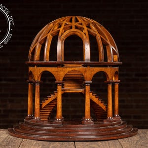 May include: A detailed wooden architectural model with a domed roof and arched openings. The model features staircases, columns, and multiple levels, all crafted from dark wood. The base is circular, and the piece is set against a dark background.