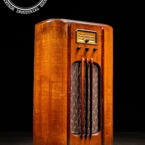 May include: Vintage wooden radio with a warm, amber finish. The radio features a speaker grill with a patterned fabric and a control panel with knobs and a dial. The cabinet has a rounded top and a rectangular shape, with the text "INDUSTRIALARTIFACTS.NET" in a circle.