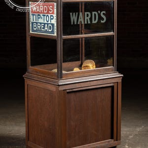 May include: Vintage wooden display case with glass panels, featuring the text "WARD'S TIP-TOP BREAD". The case has a sliding door and a lower cabinet. Inside, loaves of bread are visible. The case is made of dark wood.