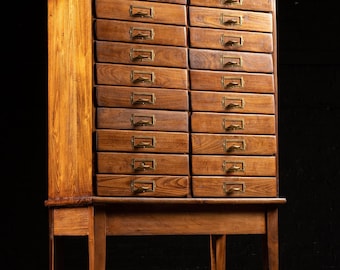 May include: A tall, vintage wooden cabinet with numerous drawers. The cabinet is made of a warm-toned wood and features brass drawer pulls. The cabinet stands on four slender legs, and the background is dark.