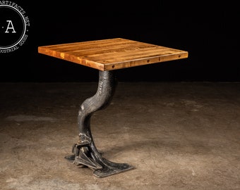 May include: A square wooden table top with a black metal base shaped like a curved leg. The table top is made of multiple wood planks with a natural wood finish. The base is made of cast iron and has a black paint finish.