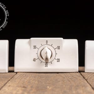 May include: A vintage white ceramic kitchen timer with a dial, flanked by salt and pepper shakers. The timer has a black and white face with numbers. The salt shaker is marked with an "S", and the pepper shaker with a "P".