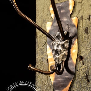 May include: A rustic, hand-forged metal hook with three hooks, mounted on a weathered wooden board. The hook is shaped like an arrowhead and has a hammered metal design. The hook is a vintage industrial style and is perfect for hanging coats, hats, or other items.