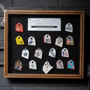 May include: A framed display of vintage advertising signs, each with a unique design and color scheme. The signs are attached to a black background and framed in wood. The text on the signs is legible, and the overall aesthetic is nostalgic.