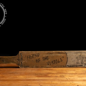 May include: A wooden paddle with the words "Friend of the Pistols" painted on it. The paddle is painted in shades of brown and gray. A stick figure drawing of a person is on the right side of the paddle.