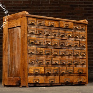 May include: Vintage wooden cabinet with numerous small drawers, each with a metal pull. The cabinet is made of light brown wood and has a rectangular shape. The cabinet is likely used for storage and organization.