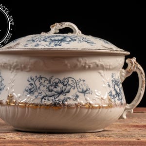 May include: Antique white porcelain chamber pot with lid, featuring blue floral patterns and gold trim. The pot has a handle and a decorative lid with a small handle. The pot is sitting on a wooden surface.