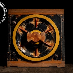 May include: A vintage industrial artifact featuring a square wooden frame with a circular design at the center. The design includes a yellow ring, black background, and a wooden center with radial spokes. The logo "IA" is visible.