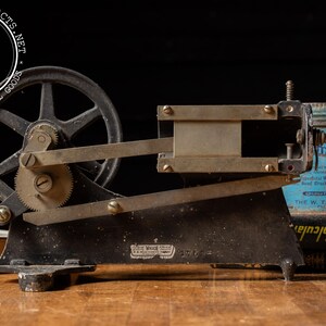 May include: A vintage industrial machine with a large gear and a metal arm. The machine is made of black metal and has a silver-colored gear. The machine is sitting on a wooden table.
