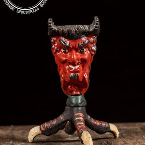 May include: A vintage cast iron devil figurine with a red face, horns, and a green and black base. The figurine has a detailed face with an angry expression and is supported by three clawed feet. The piece is likely a decorative item.