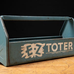 May include: A vintage blue metal tool tote with a central handle and open top. The side features the white text E-Z TOTER with a stylized graphic background. The metal shows signs of wear and age, with a rectangular base and angled sides.