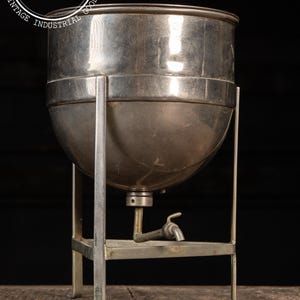 May include: Vintage silver-toned metal beverage dispenser with a spigot. The dispenser has a rounded body and sits on a rectangular stand with four legs. The top of the dispenser has a lid. The background is dark.