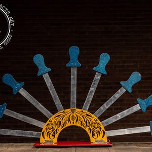 May include: A vintage stage prop featuring a fan of silver blades with blue handles. The blades radiate from a yellow, semi-circular base with a black floral design, set on a red base. The logo "INDUSTRIALARTIFACTS.NET" is in the upper left corner.