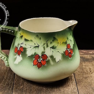 May include: Vintage ceramic pitcher with a green and white gradient, adorned with red berry clusters and leaf designs. The pitcher has a spout and a detailed handle. The overall aesthetic is reminiscent of antique tableware.
