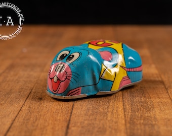 May include: A blue, yellow, and pink wind-up toy mouse. The mouse has a cartoon face with large eyes and a pink nose. The toy is made of metal and has a key on the bottom to wind it up.